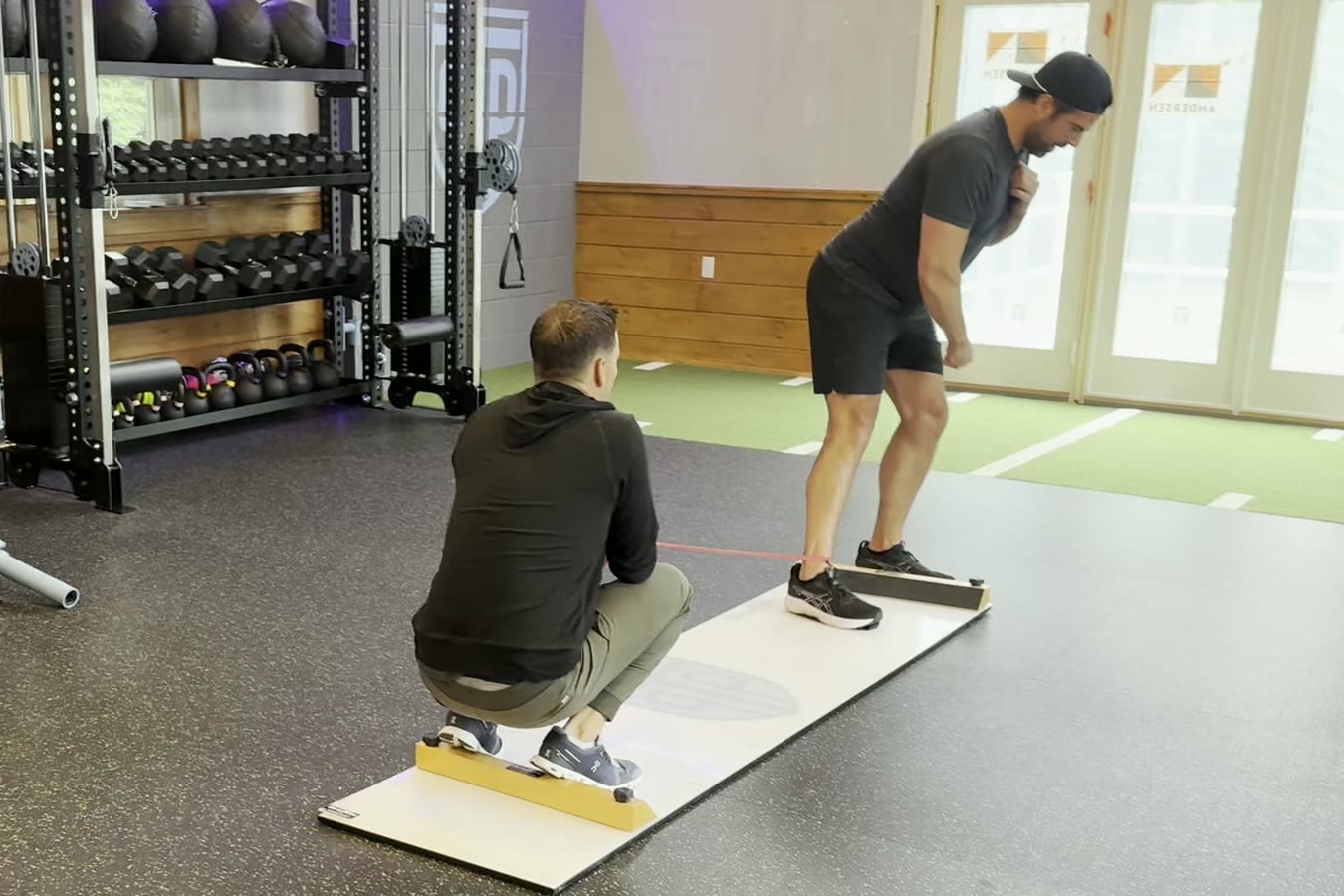 A PT Progression clinician crouches beside a client on the slide board, watching how he moves, with the turf strip behind them