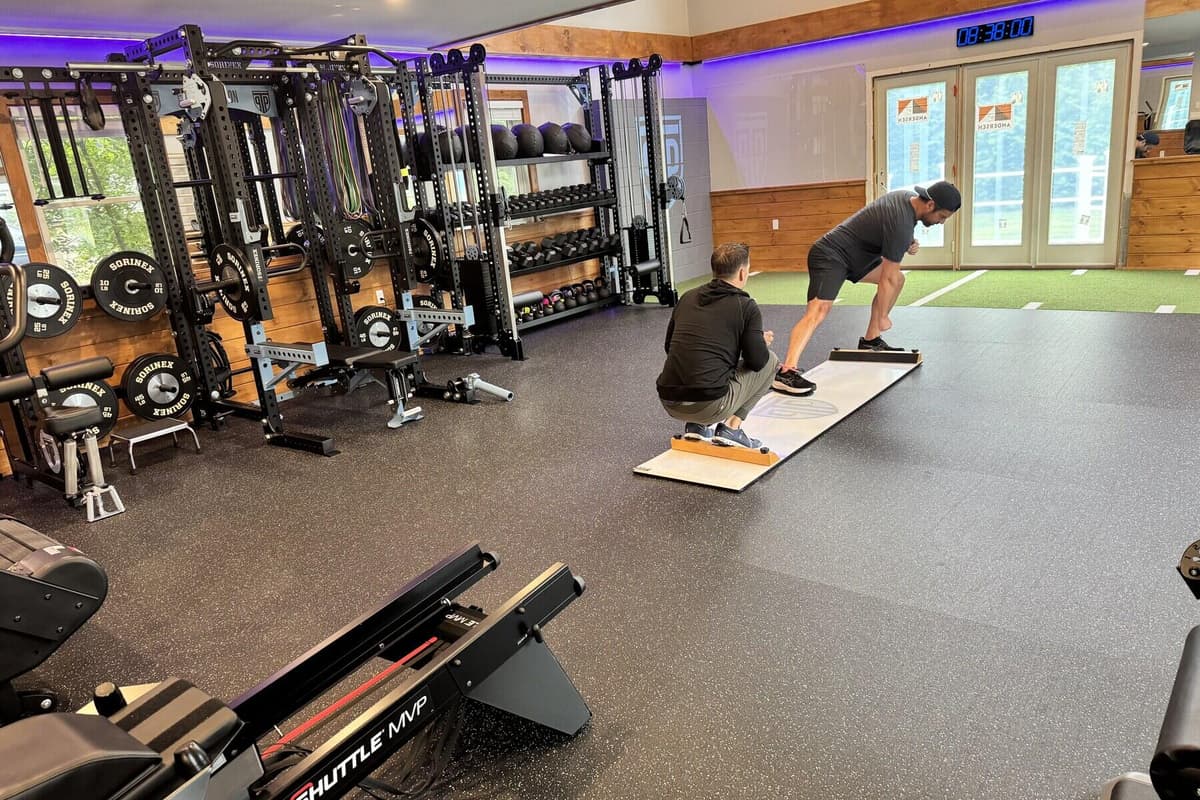 A PT Progression clinician kneels beside a client on a slide board on the training floor, with the squat racks and turf strip behind them